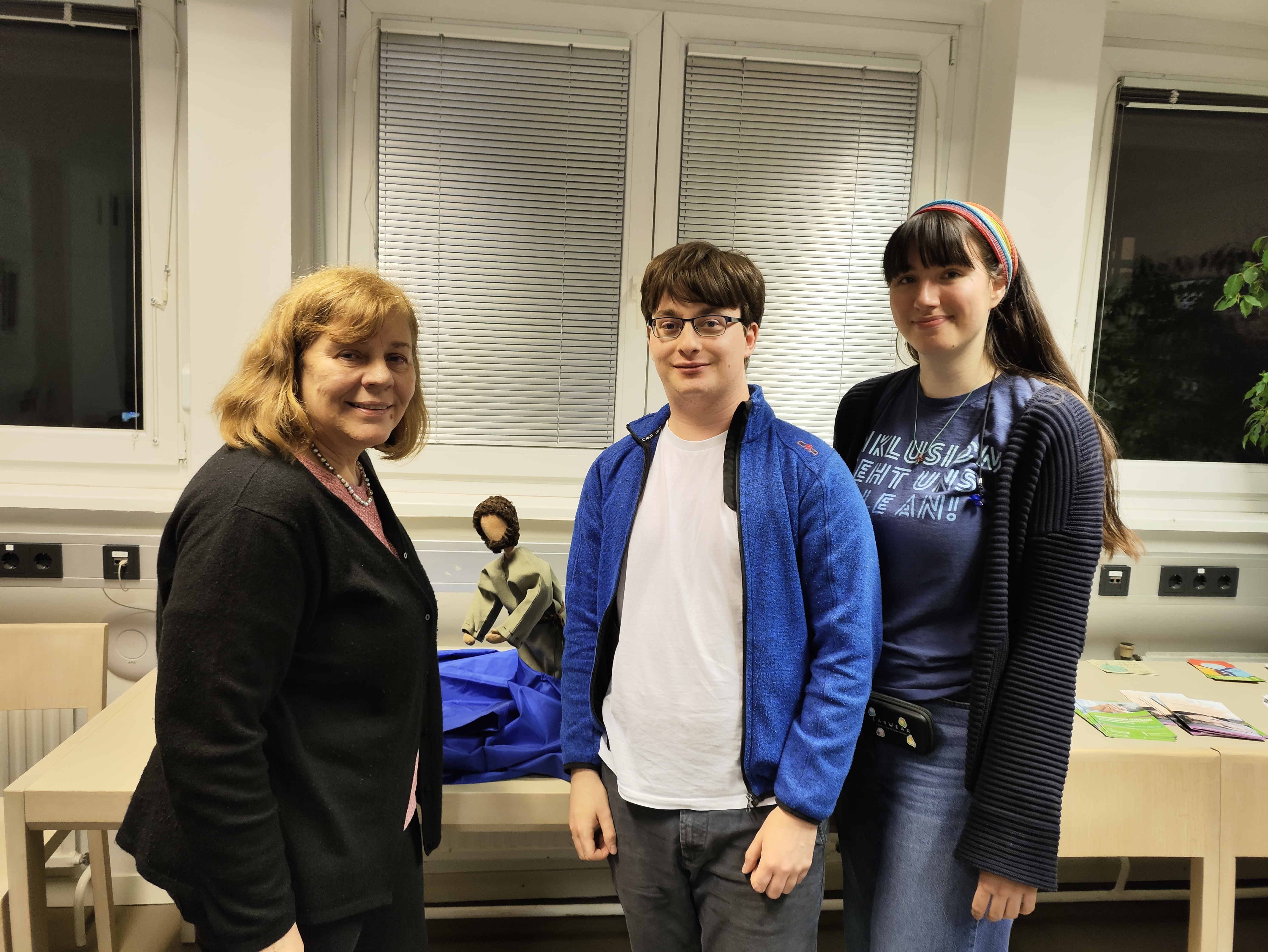 Auf dem Bild sind zwei weibliche Personen am äußeren Rand und eine männliche Person in der Mitte zu sehen. Ebenfalls sieht man im Hintergrund eine biblische Erzählfigur. 